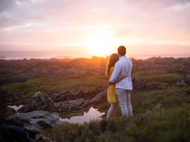 Uma mulher se perguntando como os homens apaixonados agem, abraçando o namorado enquanto assistem ao pôr do sol.