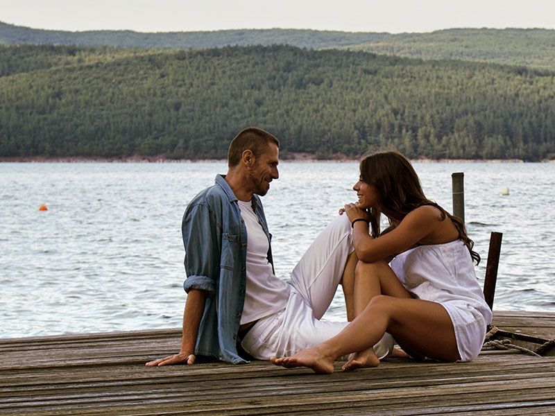 Um casal que aprendeu o que fazer no primeiro encontro, inclinando-se e conversando no cais à beira de um lago.